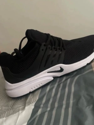 Svart vita Nike prestos  - Snygga svarta Nike sneakers med ventilerande mesh-överdel och vit, chunky sula. Skorna har svarta snören och detaljer i plast längs sidorna. Perfekta för en sportig och modern stil.