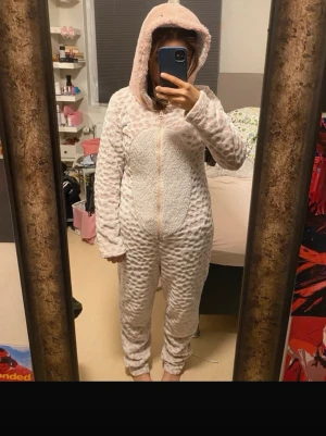 Mysig dinosaurie onesie med huva - Supergosig onesie dinosaurie pyjamas i ljusrosa och vit. Huvan har söta horn och ansikte, perfekt för chillkvällar hemma. Dragkedja framtill och långa ärmar och ben. Materialet är fluffigt och mjukt, perfekt för att mysa i.