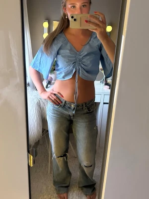 Blå croppad topp med snörning - Snygg ljusblå croppad topp med v-ringning och snörning framtill. Toppen har vida, korta ärmar och är tillverkad i ett lätt material som passar perfekt till jeans eller kjol. Trendig och ungdomlig stil med coola detaljer framtill.
