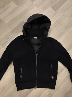 Moncler cardigan - Snygg svart pufferjacka från Moncler med vadderad huva och rygg, samt stickade ärmar och sidor. Jackan har två dragkedjeförsedda fickor framtill och ribbade muddar. 