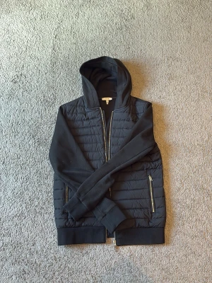 Svart hoodie från Reiss med dragkedja - Snygg svart hoodie från Reiss i storlek XS. Jackan har quiltad front, mjuka ärmar och huva samt två fickor med dragkedja. Perfekt för dig som gillar en stilren look med sportig touch. Materialet är en mix av bomull och syntet.