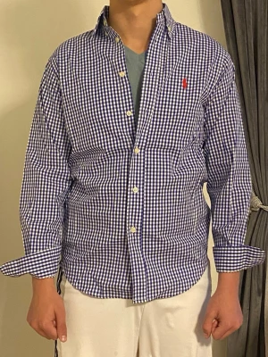 Rutig skjorta från Ralph Lauren - Snygg långärmad skjorta från Ralph Lauren i blåvitt rutigt mönster. Skjortan har klassisk krage, knäppning framtill och en röd broderad logga på bröstet. Tillverkad i mjuk bomull och passar perfekt till en avslappnad men stilren look.