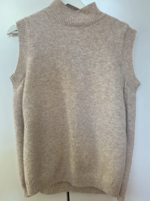 Beige stickad väst med polokrage - Mysig beige stickad väst med hög polokrage och ribbade kanter vid ärmhål och nederkant. Perfekt att styla över en skjorta eller t-shirt för en trendig lager-på-lager-look. Västen har en rak passform och är gjord i ett mjukt material.