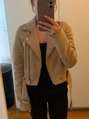 Beige mockajacka med dragkedja - Snygg beige jacka i mockaliknande material. Jackan har bälte i midjan och silverfärgade detaljer. Använd enstaka gånger.