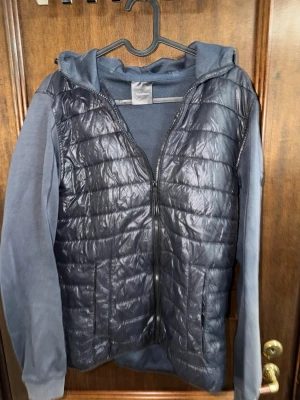 Mörkblå quiltad hoodie-cardigan - Snygg mörkblå cardiganjacka med quiltat, glansigt material på framsidan samt mjuka, ärmar och huva. Jackan har dragkedja framtill, två fickor och en sportig look. Perfekt för lager-på-lager och streetwear-vibe.