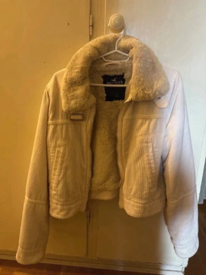 Vit pälsjacka från Hollister - Säljer en supermjuk och fluffig vit pälsjacka från Hollister. Jackan har bred krage, dragkedja framtill och är fodrad med fuskpäls både inuti och på kragen. Utsidan är i ribbad manchester och modellen är kortare i midjan. Perfekt för kalla vinterdagar!