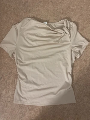 Beige topp med twistad detalj - Snygg beige topp med korta ärmar och unik twistad detalj vid halsen. Toppens material känns mjukt och stretchigt, perfekt för en trendig och stilren look. Passformen är normal och toppen är enkel att matcha med olika outfits.