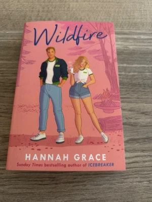 Wildfire av Hannah Grace - En romantisk skönlitterär bok med rosa omslag och illustration av två unga vuxna framför en lägereld. Boken är skriven av Hannah Grace och handlar om Russ och Aurora som möts på ett sommarläger. Perfekt för dig som gillar contemporary romance och ungdomsdrama.