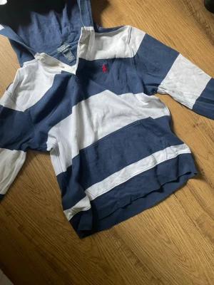 Randig hoodie med huva från Polo Ralph Lauren - Snygg hoodie med breda marinblå och vita ränder samt röd broderad logga på bröstet. Tröjan har huva, känguruficka och långa ärmar. Perfekt för en avslappnad archive stil. Tillverkad i mjukt bomullsmaterial.