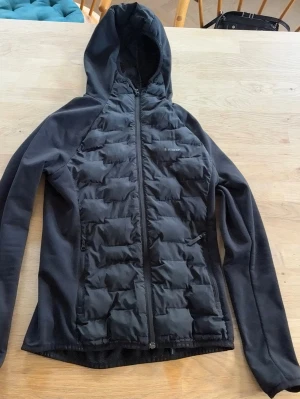 Svart peak performance vindjacka  - Snygg svart vindjacka med vadderade partier framtill och på ryggen. Jackan har huva, dragkedja hela vägen och två sidofickor med dragkedja. Materialet är en mix av slät och quiltad polyester, vilket ger en sportig look. Perfekt för blåsiga dagar och outdoor-aktiviteter.
