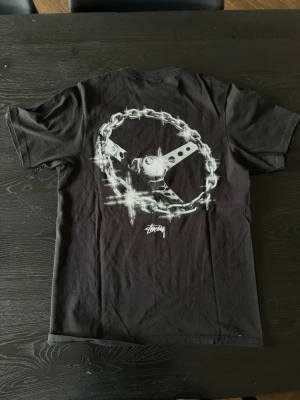 Svart Stüssy 8-ball t-shirt med kedjetryck - Svart t-shirt från Stüssy med ett stort tryck på ryggen som föreställer en glänsande ratt i kedja och en 8-ball. Perfekt för dig som gillar streetwear och coola prints.