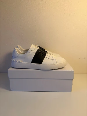 Valentino vita sneakers med svart detalj - Snygga vita sneakers från Valentino med en bred svart läderdetalj över mitten. Skorna har klassisk låg profil, vita snören och diskreta nitar på hälen. Tillverkade i läder för en lyxig känsla och stilren look.