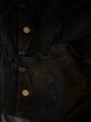 Svarta jeans  - Säljer ett par svarta jeans med raka ben och stora fickor och silverfärgade knappar framtill. Jeansen har klassisk femficksdesign och är gjorda i denim. Perfekta för en cool och avslappnad stil.