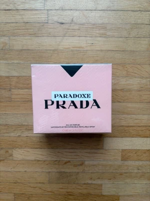Oöppnad Prada Paradoxe Eau de Parfum 90ml - Prada Paradoxe Eau de Parfum i en elegant ljusrosa kartong med svart detalj. Flaskan rymmer 90 ml och är en påfyllningsbar spray. Förpackningen har en modern och stilren design som känns lyxig och trendig. QR koden leder dig till pradabeauty’s Instagram.