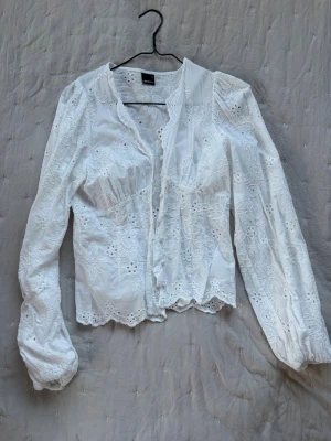 Vit broderad blus med spetsdetaljer - Superfin vit blus med broderade blommor och spetsdetaljer. Blusen har långärmade puffärmar, knäppning framtill och vågig kant nertill. Tillverkad i ett tunt, luftigt material som känns romantiskt och somrigt. Perfekt till jeans eller kjol för en söt look.