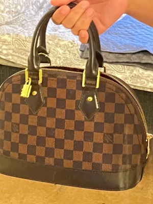 Brun handväska med rutigt mönster - Snygg handväska i brunt med klassiskt rutigt mönster och svarta detaljer. Väskan har två stadiga handtag, guldfärgade metalldetaljer och dragkedja upptill. Materialet ser ut att vara en mix av läder och canvas. Perfekt för dig som gillar tidlös och elegant stil.