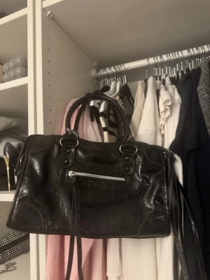 Svart väska  - Snygg svart handväska i glansigt skinnliknande material med dubbla handtag och en framficka med silverfärgad dragkedja. Väskan har dekorativa sömmar och nitar, samt långa remmar som hänger från dragkedjan. 