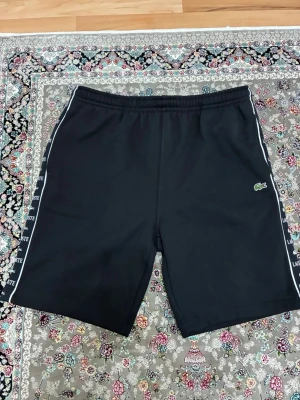 Svarta shorts från Lacoste med logga - Svarta shorts från Lacoste med elastisk midja och vita sidoremsor med Lacoste-tryck. Klassisk krokodillogga på vänster ben. Perfekta för en sportig look och sköna att bära till vardags. Materialet känns mjukt och lätt.