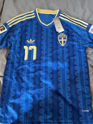 Blå Sverige matchtröja Adidas - Säljer en blå Sverige matchtröja från Adidas med gula detaljer och nummer 17 på bröstet. Tröjan har gula ränder på axlarna, Sveriges landslagsemblem och Climacool-material för bra ventilation. Snygg retroinspirerad design med mönster i tyget.