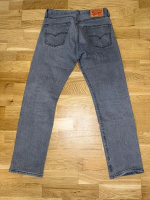 Grå raka jeans från Levi's 501 - Säljer ett par klassiska grå Levi's 501 jeans med rak passform och fem fickor. Jeansen har en ikonisk läderpatch bak i midjan och den röda Levi's-taggen på bakfickan. Tillverkade i slitstark denim, perfekta för en avslappnad och tidlös stil.