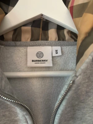 Grå zip hoodie från Burberry - Snygg grå zip hoodie från Burberry med klassiskt rutigt foder i huvan. Stl M men liten i storlek 