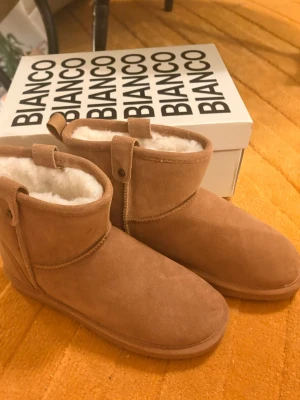 Beiga fårskinnsboots från Bianco - Mysiga och varma boots från Bianco i beige mocka med mjukt vitt foder i fårskinn. Klassisk rund tå och låg skaftlängd, perfekta för kalla dagar. Snygg enkel design med dekorativa sömmar och små flikar upptill.