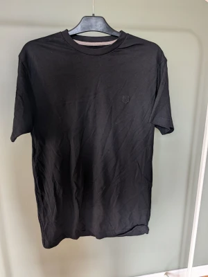 Svart t-shirt med broderad detalj - En stilren svart t-shirt i mjuk bomull med korta ärmar och rund hals. På bröstet finns en diskret broderad detalj i samma färg som tröjan. Perfekt basplagg för alla tillfällen och enkel att matcha med andra plagg.