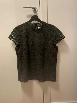Svart spetsblus med korta ärmar - Snygg svart blus i spets med korta ärmar och hög hals. Blusen har ett vackert virkat mönster över hela plagget och knäppning bak i nacken. Perfekt för dig som gillar detaljer och en elegant look.