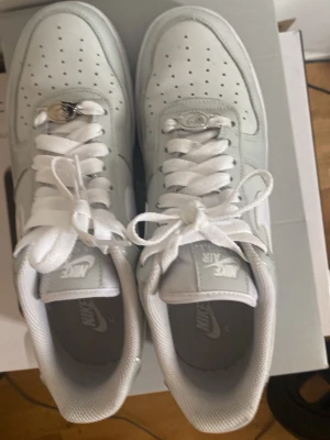 Vita Nike Air Force 1 sneakers - Klassiska vita Nike Air Force 1 sneakers med lågt skaft. Skorna har perforerad tå, vita skosnören och den ikoniska swoosh-loggan på sidan. Ovandelen är i slätt läder och sulan är vit med AIR-detalj. Perfekta för en clean och stilren look.