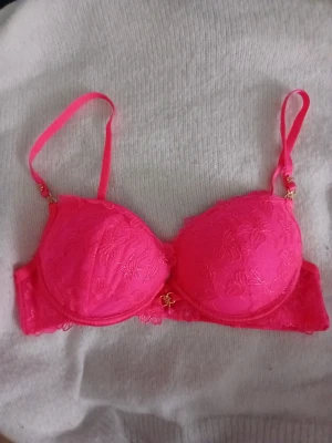 Neonrosa push-up bh med spets - Säljer då jag ska köpa i en annan storlek🥰 Säljer en neonrosa push-up bh med broderad spets och justerbara axelband. Bh:n har vadderade kupor och dekorativa gulddetaljer framtill. Materialet är en kombination av spets och slät textil för en snygg look.