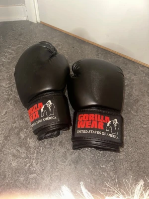 Svarta boxningshandskar Gorilla Wear - Säljer ett par svarta boxningshandskar från Gorilla Wear. Handskarna har en klassisk form med bred kardborrestängning och logga i rött och vitt på mudden. Tillverkade i slitstarkt konstläder, perfekta för träning och sparring. 6oz