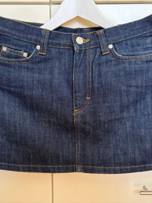 Vintage 2004 mörkblå kort jeanskjol med fickor - Snygg kort mörkblå jeanskjol i klassisk modell med fem fickor, bälteshällor och synliga kontrastsömmar. Kjolen har knapp och dragkedja framtill och är i perfekt skick. Vintage 2004 från Divided