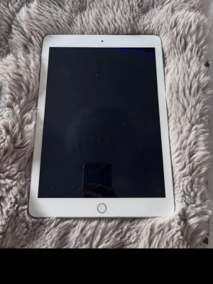 iPad med silverfärgad baksida - Generation 6 Säljer en iPad med vit ram runt skärmen och silverfärgad baksida. Plattan har en rund hemknapp och Apples logga på baksidan. Tillverkad i metall och glas, perfekt för surf, spel och streaming.