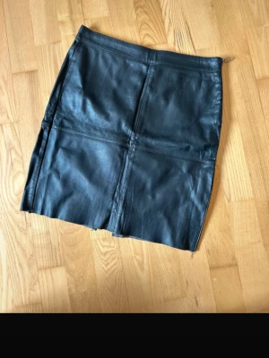 Svart skinnkjol med slits - Snygg svart kort skinnkjol med markerade sömmar och en diskret slits framtill. Kjolen har en klassisk rak passform och dragkedja i sidan. Perfekt för dig som vill ha en edgy och stilren look.