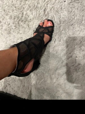 Svarta mesh-pumps med klack - Säljer ett par svarta pumps med öppen tå och chunky klack. Skorna har ett coolt mesh-tyg med dekorativa band över foten och runt ankeln. Perfekta för dig som vill ha något trendigt och edgy till din outfit.