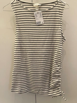 Randig ribbad linnetopp med knyt - Säljer en vit och svart ribbad linnetopp med tunna horisontella ränder och knyt i sidan. Toppen är ärmlös och har en enkel, rak passform som passar perfekt till jeans eller kjol. Superenkel att styla för en chill look.