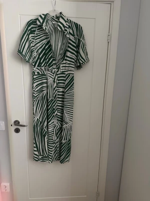 Grönvit långklänning med krage - Snygg långklänning i grönvitt randigt mönster med krage och korta ärmar. Klänningen har knappar framtill och ett avslappnat fall. Perfekt för dig som gillar statement-plagg med unik design.
