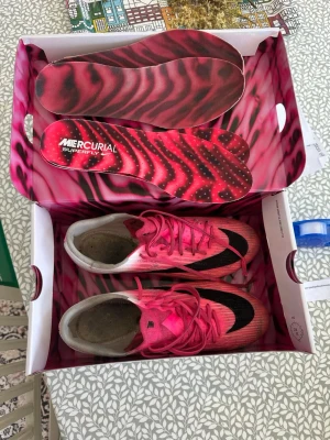 Nike Mercurial Elite Superfly rosa fotbollsskor - Säljer ett par Nike Mercurial Superfly fotbollsskor i rosa och vitt med svarta detaljer. Skorna har en låg profil, snörning och mönstrade sulor för bra grepp på planen. Innersulorna har ett unikt zebramönster i rosa och svart. Perfekta för dig som vill sticka ut på planen.