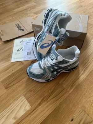 Asics  - Snygga Asics sneakers i vitt och silver med svarta detaljer. Skorna har en sportig design med mesh- och syntetmaterial för bra andningsförmåga och komfort. Klassisk Asics-logga på sidan och tungan. Perfekta för dig som gillar streetwear och sportig stil.