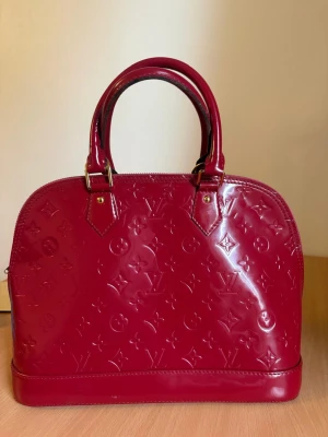 Röd Louis Vuitton Alma handväska - Snygg röd handväska från Louis Vuitton i lackat läder med präglat monogrammönster. Väskan har två handtag, guldfärgade metalldetaljer och dragkedja med LV-logga. Klassisk bågformad design som ger en lyxig känsla.