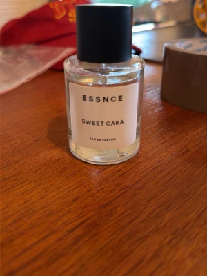 ESSNCE Sweet Cara Eau de Parfum - Minimalistisk parfymflaska från ESSNCE med doften Sweet Cara. Flaskan är genomskinlig i glas med en enkel vit etikett och svart cylinderformad kork. Modern och stilren design som passar perfekt på sminkbordet. Använd få tal gånger!!