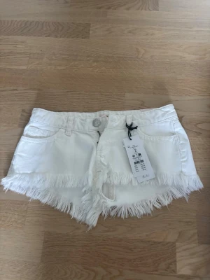Vita jeansshorts med låg midja - Säljer ett par helt nya vita shorts i jeanstyg och fransig kant från Gina 14+. Dom har lapp och allt kvar. Shortsen har klassisk femficksdesign, bälteshällor och stängs med knapp och dragkedja. Perfekta för sommaren och ger en avslappnad vibe.