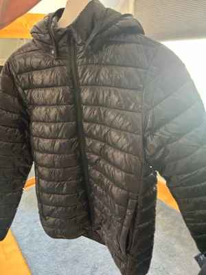 Svart quiltad vindjacka med huva - Snygg svart vindjacka med quiltad design och huva. Jackan har dragkedja framtill och två sidofickor. Tillverkad i lätt, glansigt material som ger en sportig look. Perfekt för blåsiga dagar och enkel att matcha med olika outfits.
