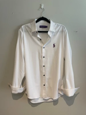 Purple Lable skjorta Ralph Lauren - Klassisk vit skjorta från Ralph Lauren med mörka knappar och lila broderad logga på bröstet. Skjortan har lång ärm, krage och rak passform. Tillverkad i mjukt bomullstyg för en stilren look.