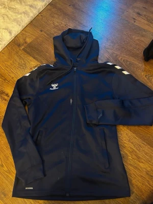 Mörkblå hoodie från Hummel - Snygg mörkblå hoodie från Hummel med dragkedja framtill och vita detaljer på axlarna. Huvan är justerbar och materialet är mjukt och sportigt, perfekt för chill eller träning. Hummel-logga på bröstet och sidofickor för småprylar. Tröjan är i jätte fin skick utöver fläckarna som syns på bilderna (går inte bort i tvätten, har dock ej testat behandla det)