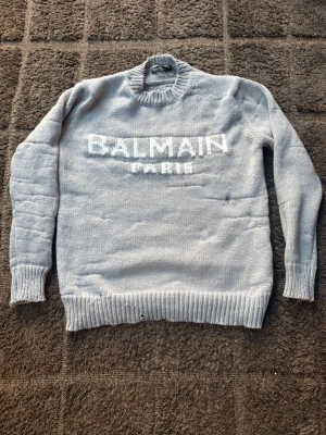 Ljusgrå stickad tröja från Balmain - Snygg ljusgrå stickad tröja från Balmain Paris med vit logga framtill. Tröjan har ribbstickad hals, ärmslut och nederkant. Perfekt för dig som vill ha en trendig och bekväm look med exklusiv känsla.