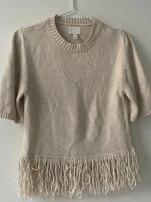 Beige stickad tröja med fransar H&M - Säljer en beige stickad tröja från H&M med korta ärmar och rund halsringning. Tröjan har ribbade muddar och coola fransdetaljer längs nederkanten som ger en trendig look. Perfekt att styla till jeans eller kjol.
