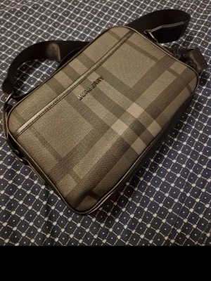 Rutigt axelväska i svart och grått - Snygg axelväska med klassiskt rutmönster i svart och grått. Väskan har justerbar axelrem i textil och dragkedja upptill. Tillverkad i tåligt material med stilren design och diskret logga på framsidan. Perfekt för dig som vill ha en trendig och praktisk väska.