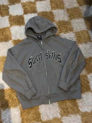 Sweet sktbs zip hoodie - Grå hoodie med dragkedja och huva, samt svartvit text 'Sweet Sktbs' i gotisk stil på bröstet. Mjuk bomullskänsla, ribbade muddar och avslappnad passform. Perfekt för en chill streetwear-look.