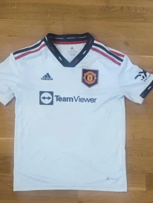 Manchester United bortatröja Adidas - Säljer en vit Manchester United bortatröja från Adidas med svarta och röda detaljer. Tröjan har klubbmärke på bröstet, Adidas-logga och TeamViewer sponsortryck. Tillverkad i funktionsmaterial, perfekt för fotbollsträning eller match.